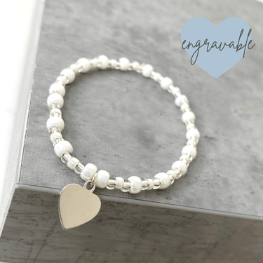 Pearl bracelet with a heart charm on a gray surface, branded 'engravable'.