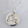 A silver round disc pendant necklace with a hand-drawn outline of Ireland, displayed on a light gray jewelry box.