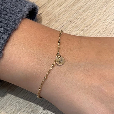 A delicate gold chain bracelet with a small round disc charm featuring a monogram, worn on a person's ankle against a light wood background.