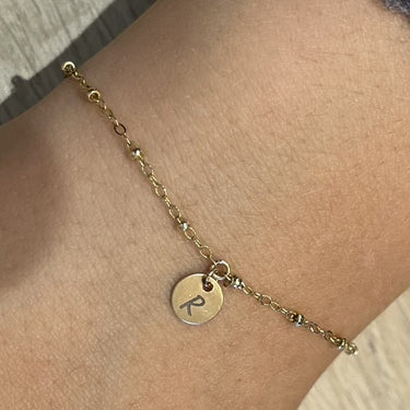 A delicate gold chain bracelet with a small round engraved disc charm, shown worn on a person's wrist with a gray knit sleeve visible.
