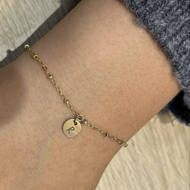 A delicate gold chain bracelet with a small round engraved disc charm, shown worn on a person's wrist with a gray knit sleeve visible.