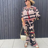 Woman wearing a colorful outfit with a patterned top and pants, holding a gold handbag against a wooden panel background.