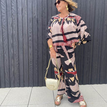 Woman wearing a colorful outfit with a patterned top and pants, holding a gold handbag against a wooden panel background.