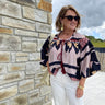 Woman wearing a patterned blouse and sunglasses standing against a stone wall.