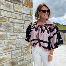 Woman wearing a patterned blouse and sunglasses standing against a stone wall.