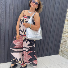 Woman wearing a colorful patterned playsuit and sunglasses, holding a white bag against a dark wall.