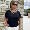 Woman wearing a black t-shirt and colorful necklace standing against a stone wall.