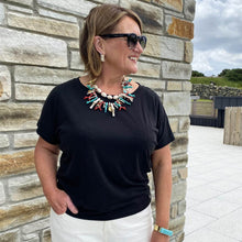 Woman wearing a black t-shirt and colorful necklace standing against a stone wall.