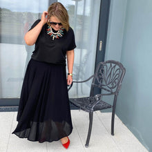 Woman wearing a black outfit with a long skirt and red shoes, standing near a chair.