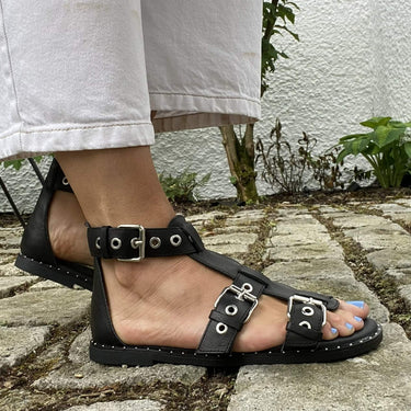 Black sandals worn on a stone pavement with a white wall and greenery in the background