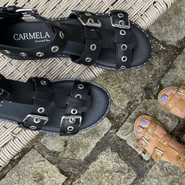 Black sandals with 'Carmela' branding on a stone pavement.