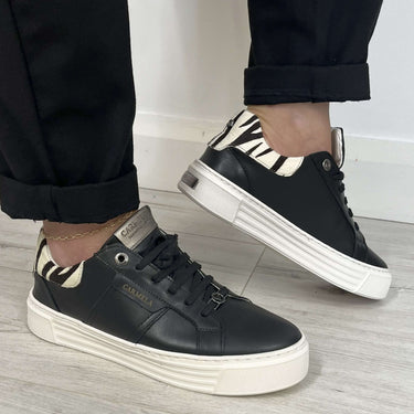 Black sneakers with white soles worn by a person on a light wooden floor.