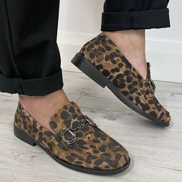 Leopard print loafers worn with black pants on a light wooden floor.