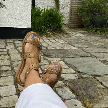 Person wearing tan sandals with white pants on a stone patio in front of a white brick building.