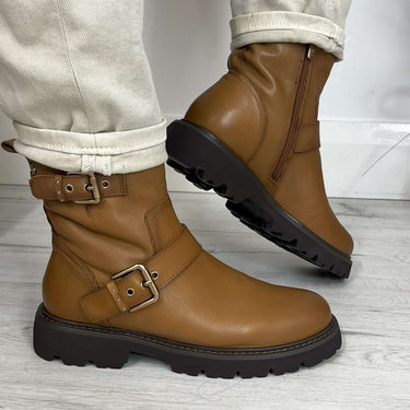 Brown leather boots with buckles worn with light-colored pants on a wooden floor.