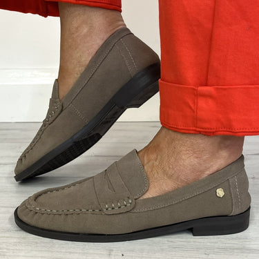 A person wearing taupe-brown suede penny loafers with black soles and bright red cuffed trousers, standing on a light gray wood floor.