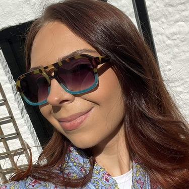 Woman wearing sunglasses and a patterned top against a white wall.