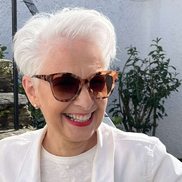 Woman wearing sunglasses and a white jacket with a red skirt in front of a building.