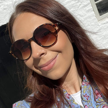 Woman wearing sunglasses and a patterned scarf against a white wall.
