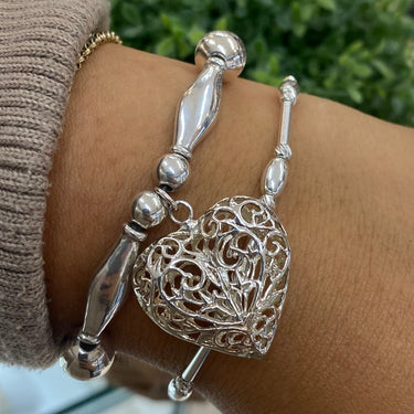 A close-up of a person's wrist wearing two silver bracelets: one beaded and one featuring an intricate filigree heart charm, set against a blurred indoor background with a green plant.