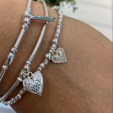 A close-up of a person's wrist wearing three silver charm bracelets, including a heart-shaped charm, a bar charm, and a beaded bracelet with a heart pendant, with a green plant in the background.