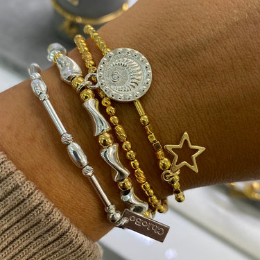 A close-up of a person's wrist wearing multiple bracelets, including a silver-toned beaded bracelet, a silver-toned disc charm bracelet, a gold-toned beaded bracelet with a star charm, and a silver-toned snake charm bracelet.