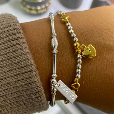A close-up of a person's wrist wearing two bracelets: a silver beaded bracelet with a small gold heart charm and a silver bar bracelet with a rectangular tag. The background shows jewelry display stands in a store.