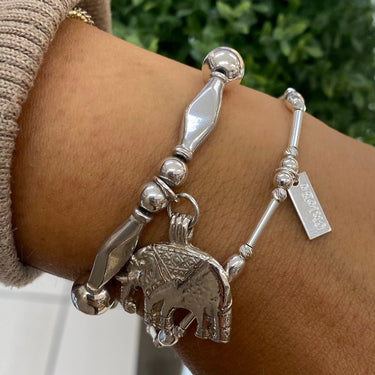 A close-up of a person's wrist wearing two silver-toned bracelets, including an elephant charm bracelet and a beaded bracelet with a small tag, against a blurred indoor background with a green plant.