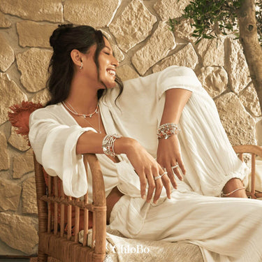 Woman in a white dress sitting on a wooden chair against a stone wall, wearing jewelry.