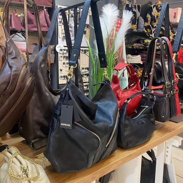 A collection of designer handbags from Emporio Armani displayed on a wooden counter in a retail store. The bags include a black leather slouchy hobo bag with a gold zipper, a dark brown leather bag, a red leather bag, and a black leather bag with a black strap.