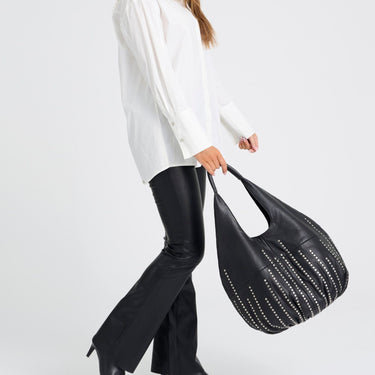 A person wearing a white blouse and black flared pants carries a large black slouchy shoulder bag with silver studs against a light gray studio background.