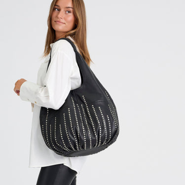 A woman wearing a white shirt and black pants carries a large black leather slouchy shoulder bag decorated with vertical silver studded stripes, set against a plain light gray background.