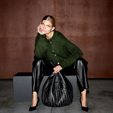 A model sits on a dark block against a textured brown wall, wearing a dark green knit cardigan, black leather pants, and black heels, while holding a large black studded handbag.