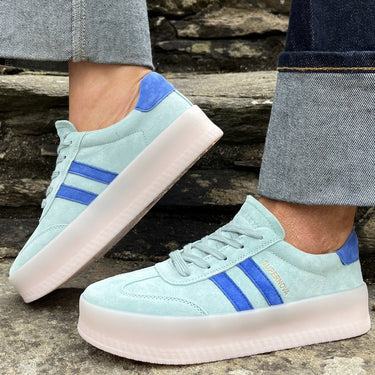 Light blue sneakers with two blue stripes worn on a stone surface