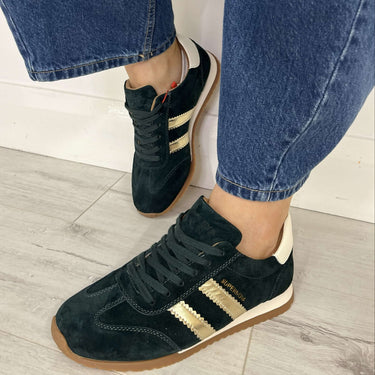 A person wearing black suede sneakers with gold stripes and blue jeans, standing on a light gray wood floor against a white wall.