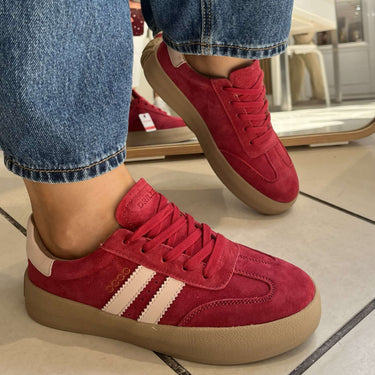 Red suede low-top sneakers with white side stripes and beige gum soles, shown on a person wearing blue jeans in a store setting.