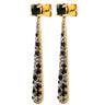 A pair of gold drop earrings with a round black gemstone at the top and a long, tapered bar studded with black and gray faceted crystals, shown on a plain white background.