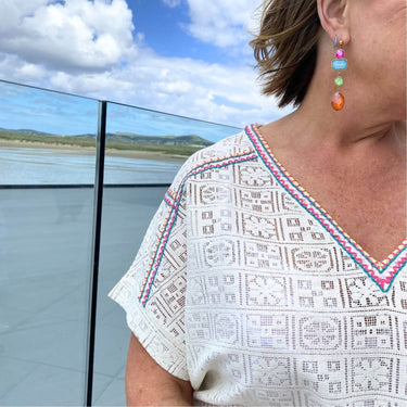 Woman wearing a white embroidered top with colorful trim, standing by a waterfront with mountains in the background.