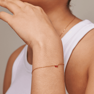 Close-up of a person wearing a gold bracelet with a red charm on a neutral background