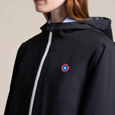 Close-up of a person wearing a black hooded jacket with a white front zipper and a small circular red, blue, and white logo patch on the left chest, against a plain light beige background.