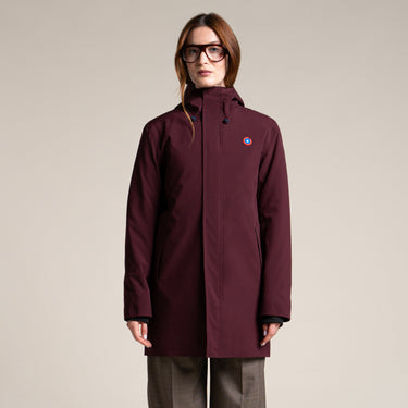 A woman modeling a burgundy hooded jacket with a small circular logo on the left chest, paired with wide-leg gray trousers and dark shoes, against a solid light beige background.