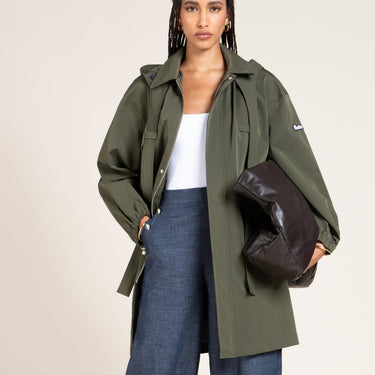 A model wearing an olive green hooded jacket over a white top and wide-leg blue jeans, holding a dark brown leather clutch bag against a plain beige studio background.