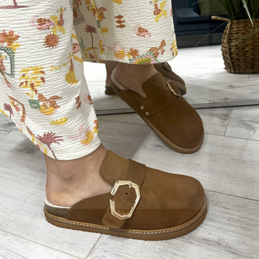 Brown shoes with gold buckles worn with floral pants on a wooden floor.