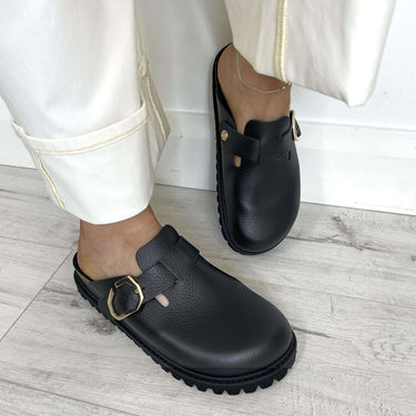 Black clogs with gold buckles worn with white pants on a light wooden floor.