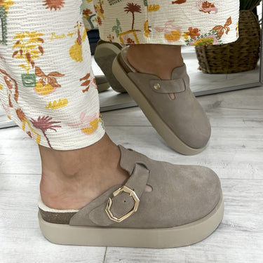 Brown shoes with a gold buckle worn by a person in floral pants on a light wooden floor.
