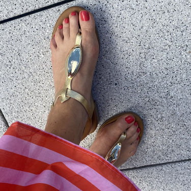 Gold sandals worn on a foot with red nail polish on a gray pavement.