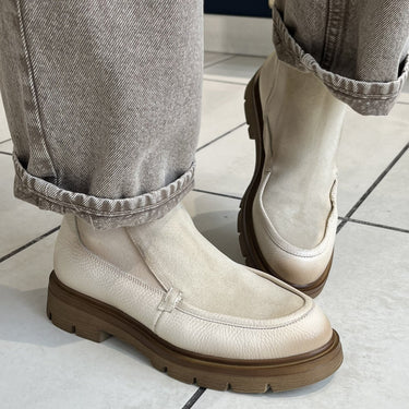 A person wearing light gray cuffed jeans and off-white slip-on loafers with brown soles, standing on a light gray tiled floor.