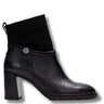 A black leather ankle boot with a chunky block heel, a black suede-like collar, and a braided strap detail accented by a silver-tone circular charm, shown against a plain white background.