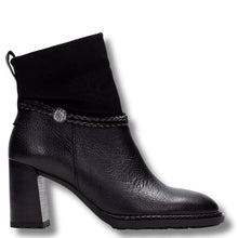 A black leather ankle boot with a chunky block heel, a black suede-like collar, and a braided strap detail accented by a silver-tone circular charm, shown against a plain white background.