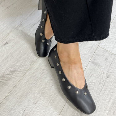 Black leather low-heel pumps with silver-tone studs, shown on a model wearing black pants against a light gray wood-look floor.